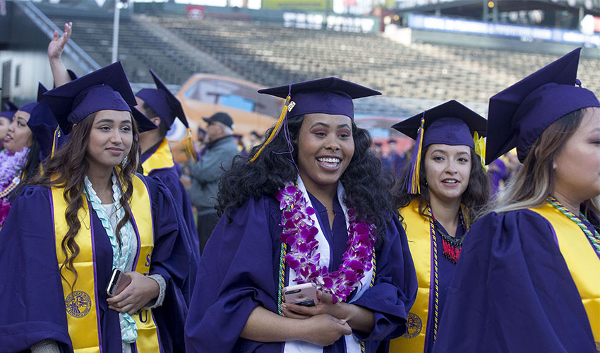 Three women dressed in SF State regalia in Oracle Park smile at the camera