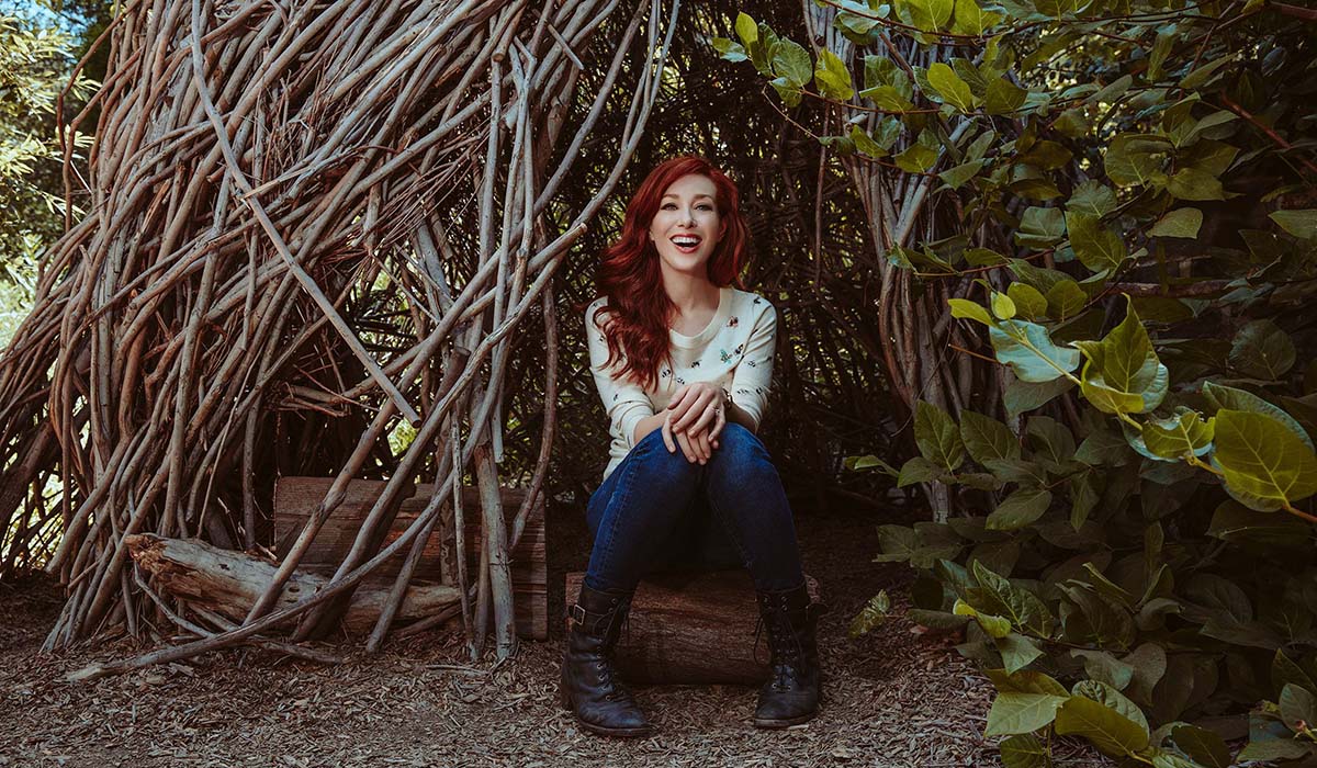Alie Ward sitting and smiling by a tree hut