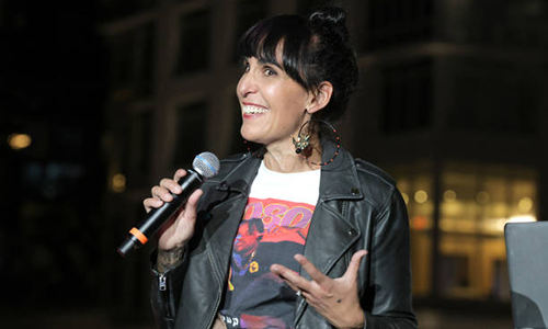 Felicia Viator holds a microphone while speaking and wearing a black leather jacket and a 4080 Magazine T-shirt