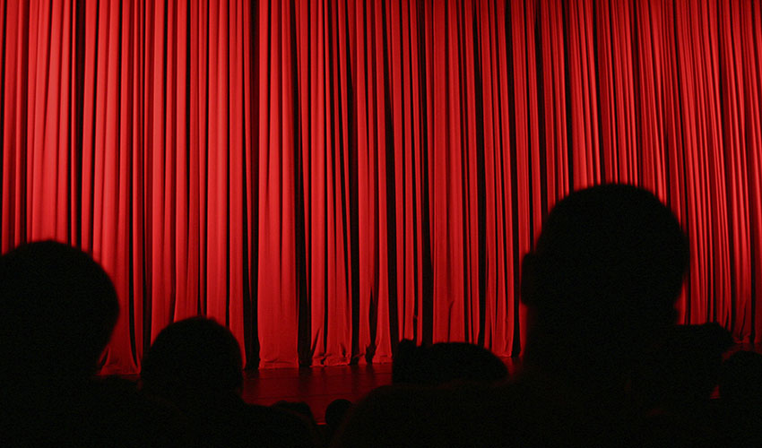 Photo of a theater stage with a red curtain and silhouettes of people facing the stage
