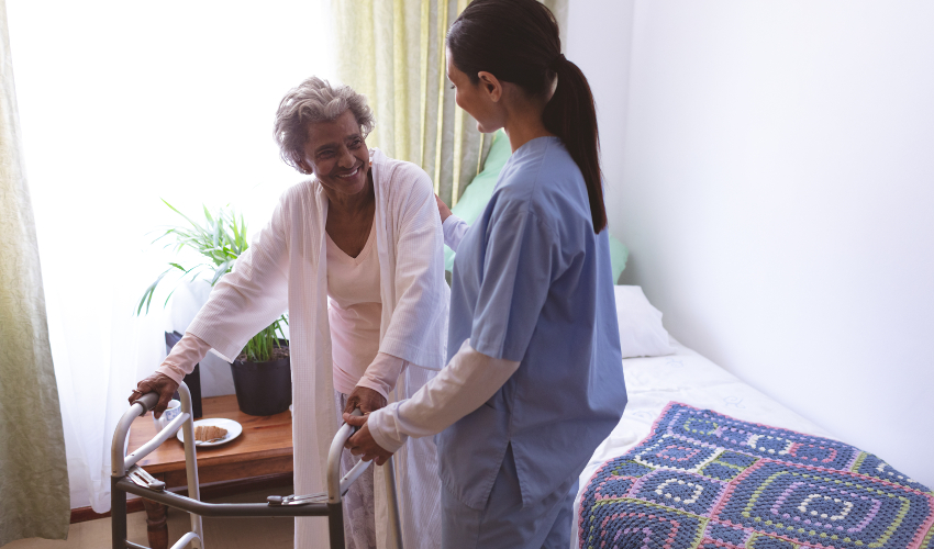 An elderly woman using walker smiles at a caretaker in a bedroom