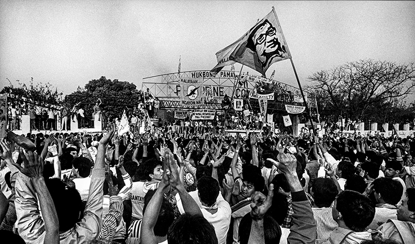 Thousands of men and women, arms raised in the air, form the letter "L" with their thumbs and forefingers to represent the word "Laban," which means "to fight" In Tagalog. A flag bearing the likeness of assassinated Filipino political leader Benigno Aquino, Jr. waves over the crowd. Photo credit: Kim Komenich for the San Francisco Examiner ©2011/ The Pictorial Collection, Bancroft Library, University of California, Berkeley.