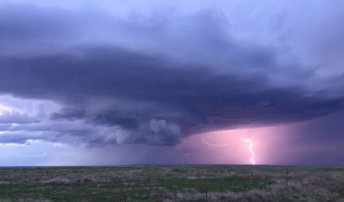 Bolts of lightning extend vertically from a large, lavender thundercloud to the grassy plains thousands of feet below.