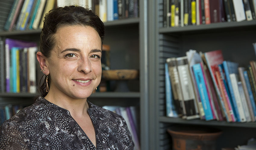 Assistant Professor Alexandra Pappas is seated in her office; the bookcase in the background is lined with books about Greek art and literature and also features a kylix, a Greek vessel used for drinking wine.