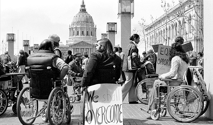 Black and white photo of protesters in wheelchairs by San Francisco City Hall with one holding sign with text We Shall Overcome