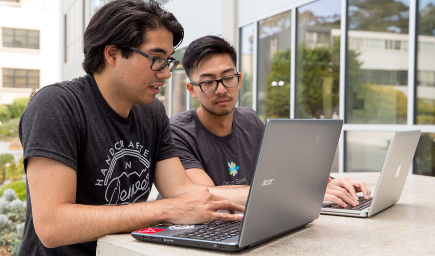 A photo of Kevin Lay and Matthew Serna working on two laptops in the SF State quad.