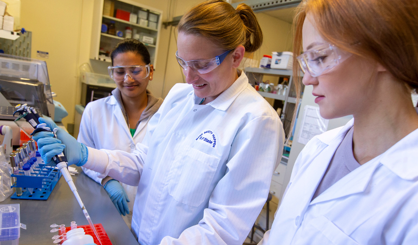 Researcher in white lab coat using a pipette while two students watch