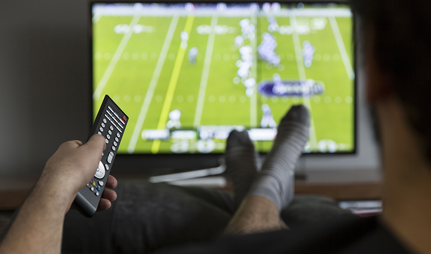 A photo of a man sitting on a couch watching football on TV.