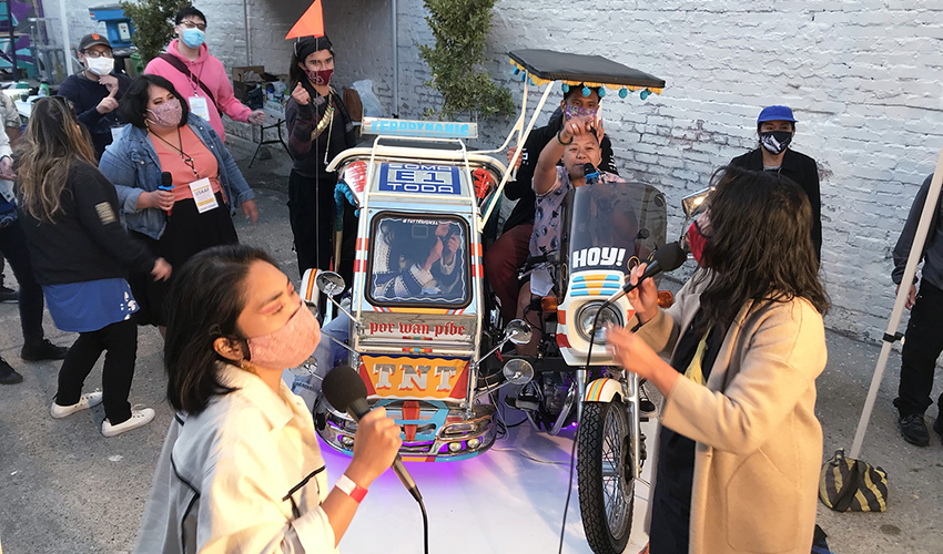 Two people sing karaoke in front of the TNT Traysikel outdoors while a small crowd watches.