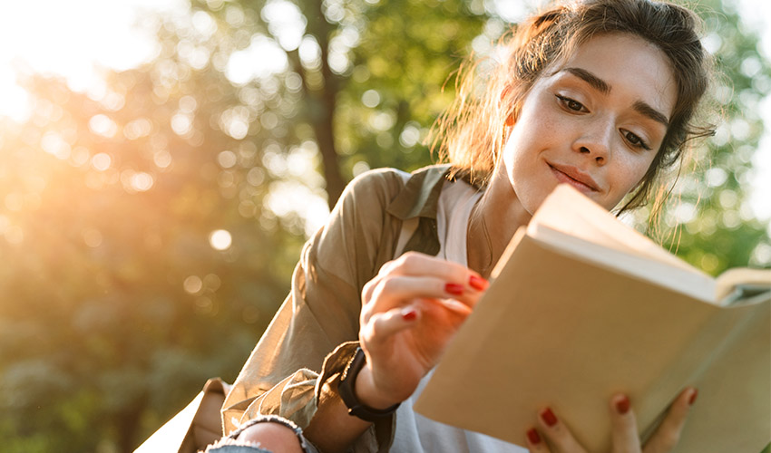 A teenage girl reading a book outside with trees surrounding her