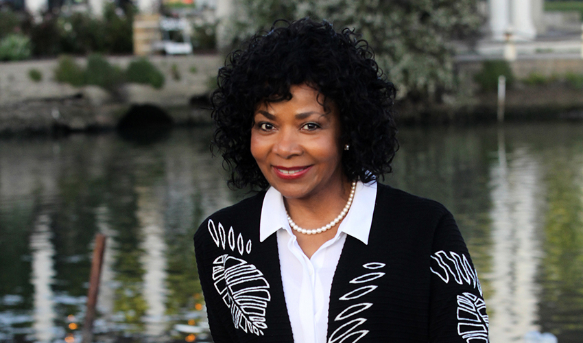 Alumna Ramona Tascoe, wearing a white blouse and black and white suit, is pictured with a lake in the background, leaning against a curved piece of driftwood.