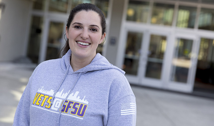 Elizabeth (Lizz) Lauren Mayhill, standing outside the J. Paul Leonard Library, wears a VETS@SFSU hoodie.