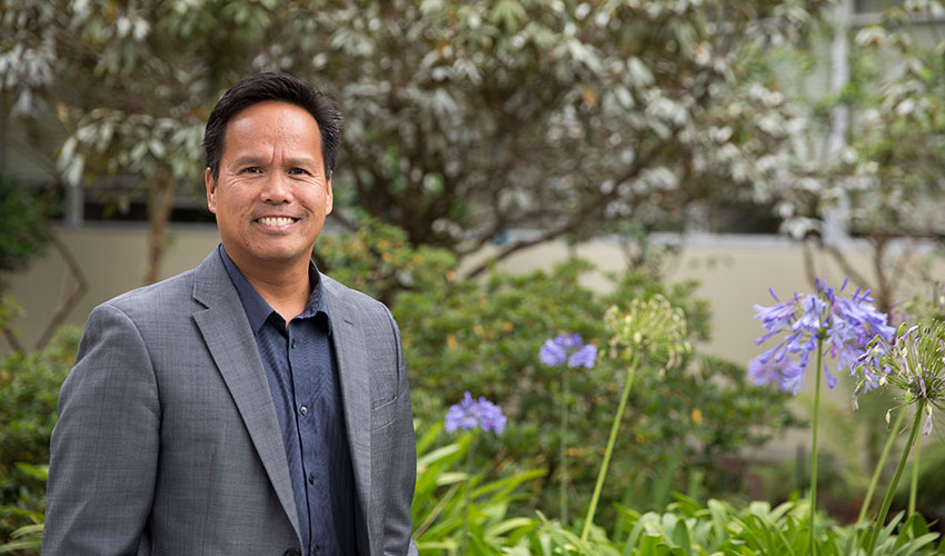 Health & Social Sciences Dean Alvin Alvarez, wearing a gray suit, stands in front of plants, trees and flowers.
