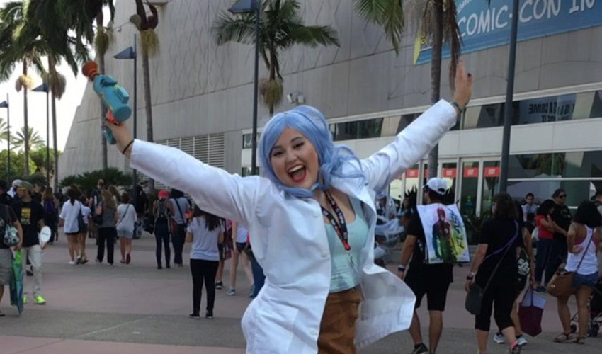zul Sanchez-Macias wears a wig and lab coat and poses with her arms outstretched.