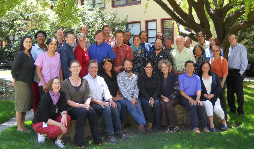 A group of about thirty faculty members smile at the camera, seated and standing, with two trees and a yellow building in the background.