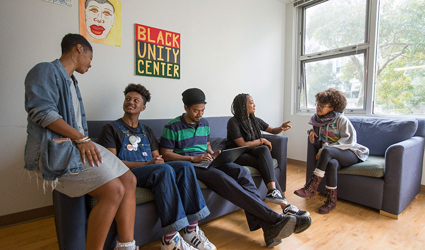 Four students sit next to a professor on a couch under a sign that reads Black Unity Center.