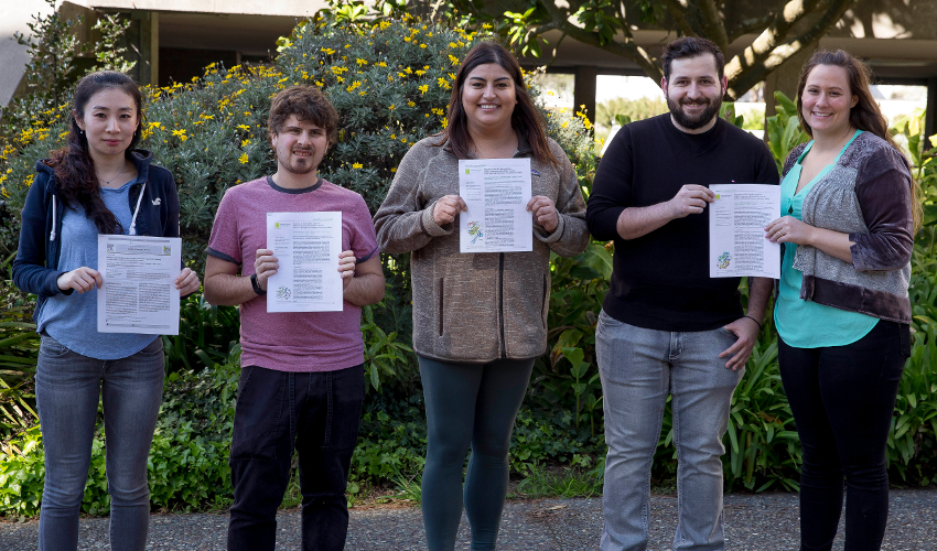 Five students holding papers smile at the camera, with green bushes in the background