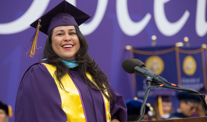 Woman in purple and yellow graduation regalia, standing at podium and smiling