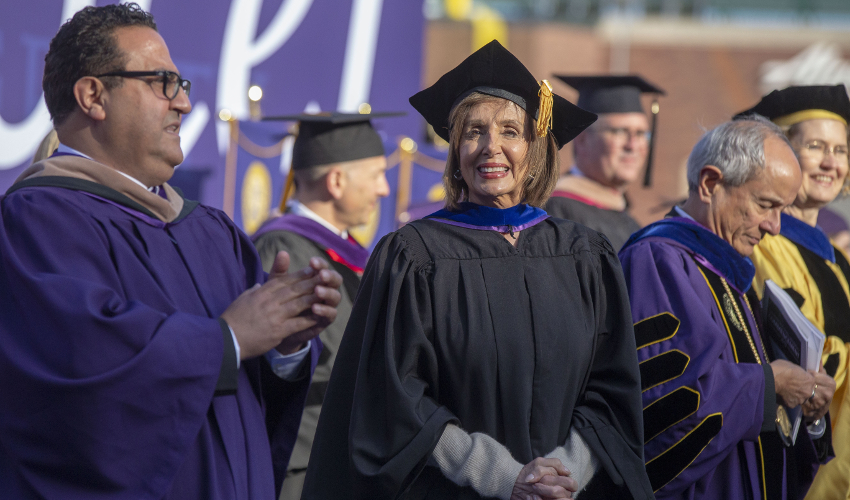 Nancy Pelosi faces the camera dressed in a black robe and cap, surrounded by others in purple.