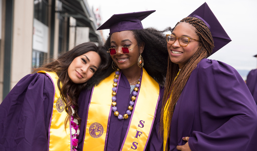 Three students in purple graduation robes and caps smile at the camera