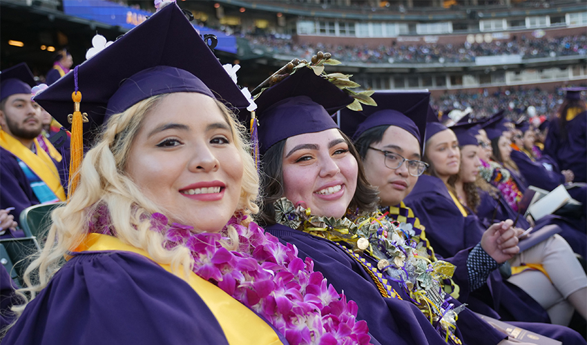 Graduate dressed in regalia sit and smile at the camera at Oracle Park