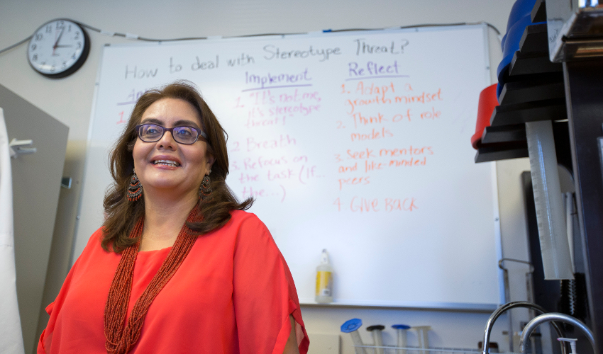 Leticia Márquez-Magaña, dressed in red, stands in front of a whiteboard with writing on it