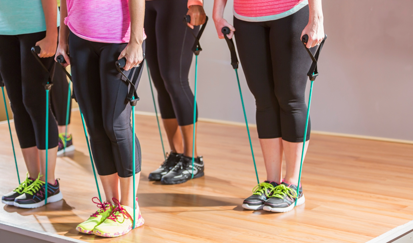 A group of women conduct resistance band exercises in a gym.