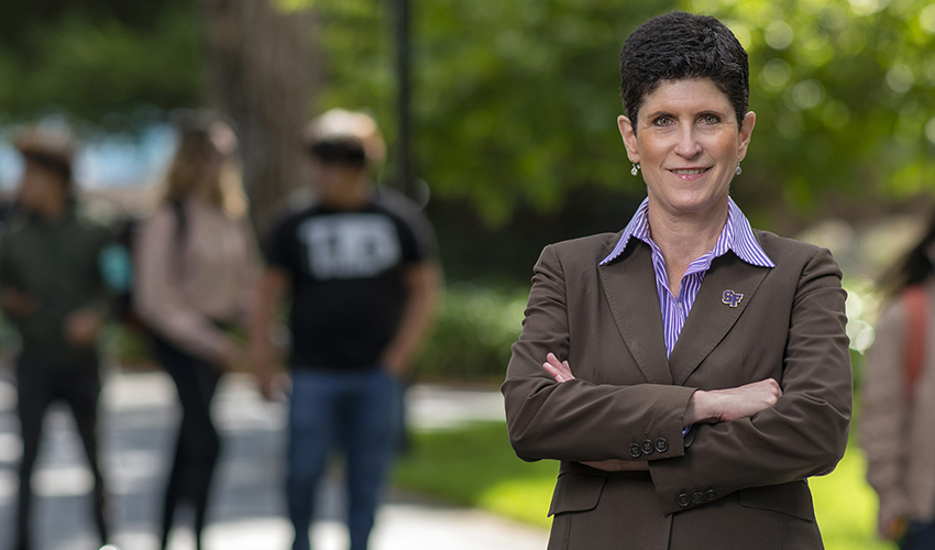 Woman in brown suit with her arms crossed standing against a background that shows green foliage and people walking.