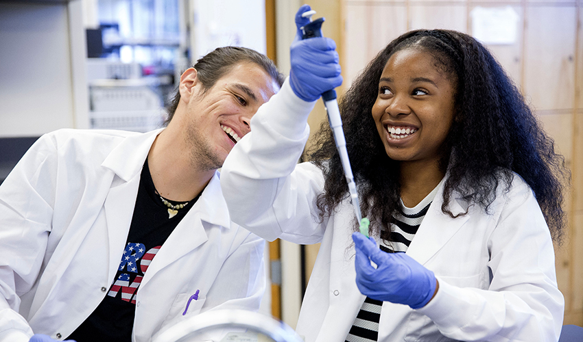 Two people side-by-side in white lab coats. Both are smiling while one is using a