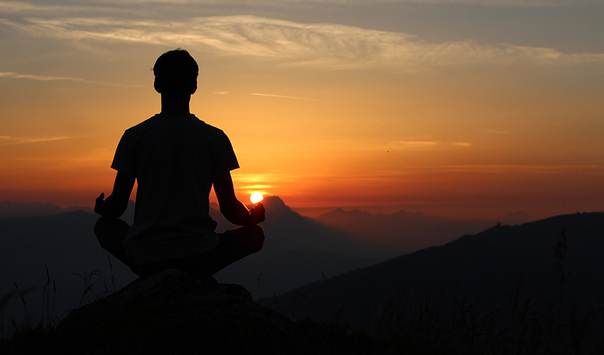 Silhouette of a person sitting – assumably meditating — while looking at a fading sun on the horizon.