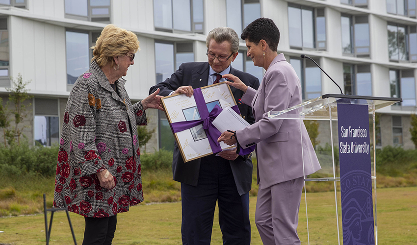 Woman in lavender suit presenting a large framed photo to two people