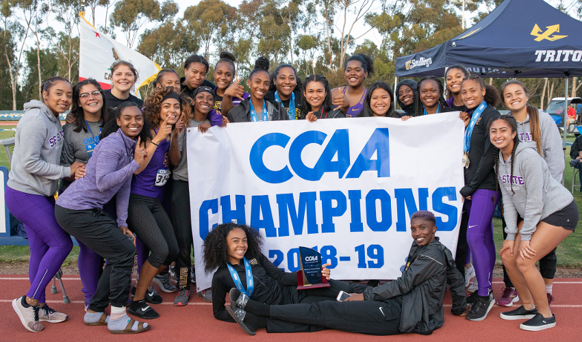 Students at a running track hold up a sign reading "CCAA Champions 2018-19"