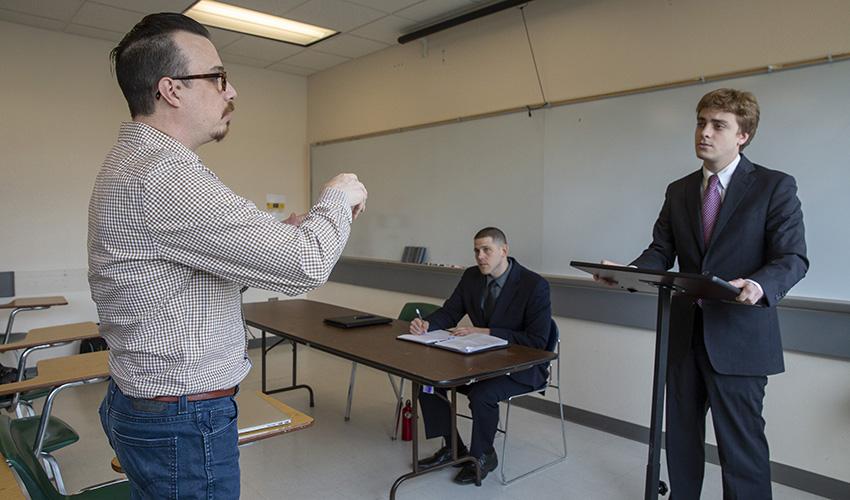 Moot Court Club president stands behind a podium in a classroom as SF State Political Science Assistant Professor Nick Conway and Aaron Gamez watches as he sits at a nearby table
