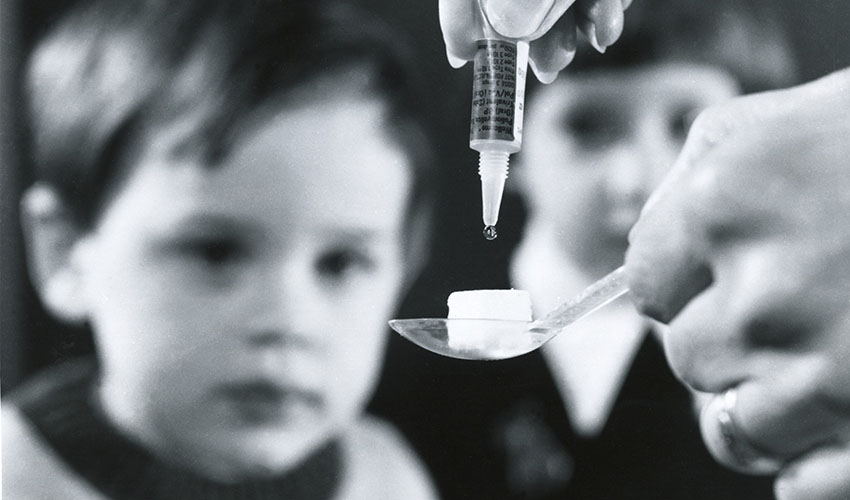Syringe drips vaccine on to a sugar cube held by a spoon as a child looks on in the background