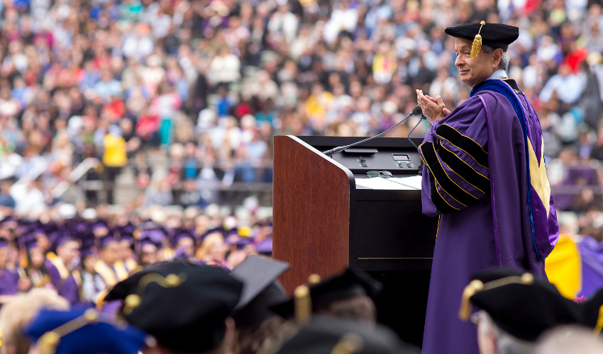 Leslie E. Wong stands at a podium in academic regalia with a crowd in the background