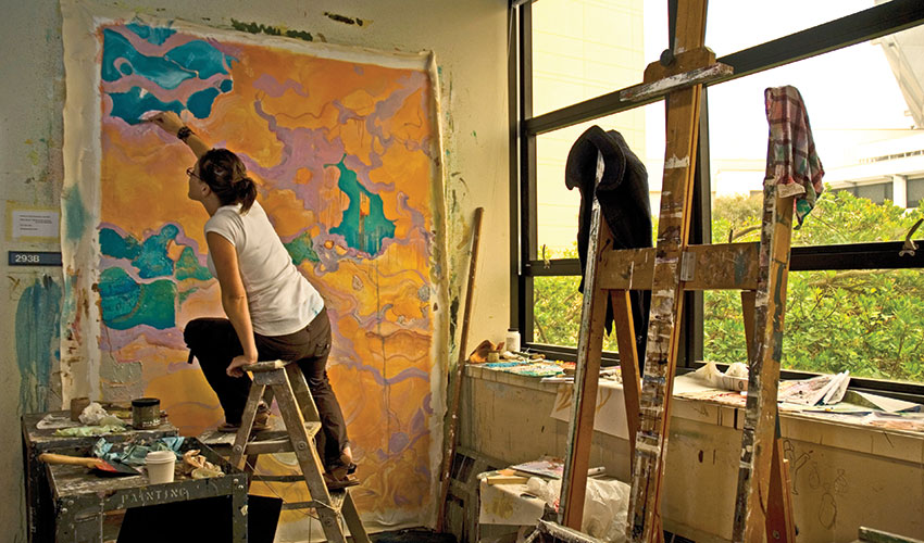 A woman sits on a ladder while working on a large orange painting.