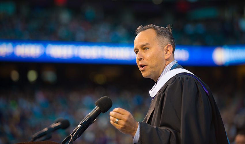 Jonas Rivera stands at a podium speaking in front of a crowd of graduates at AT&T Park