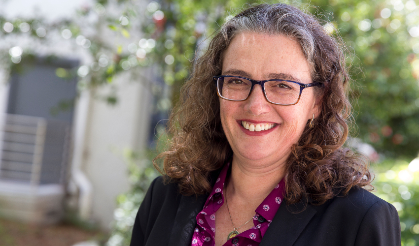 Woman in blazer and purple shirt smiles at the camera with a tree and a gray building in the background