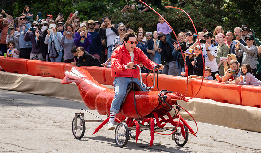 A man smiles as he rides a shrimp-car down a hills as spectators watch and cheer.