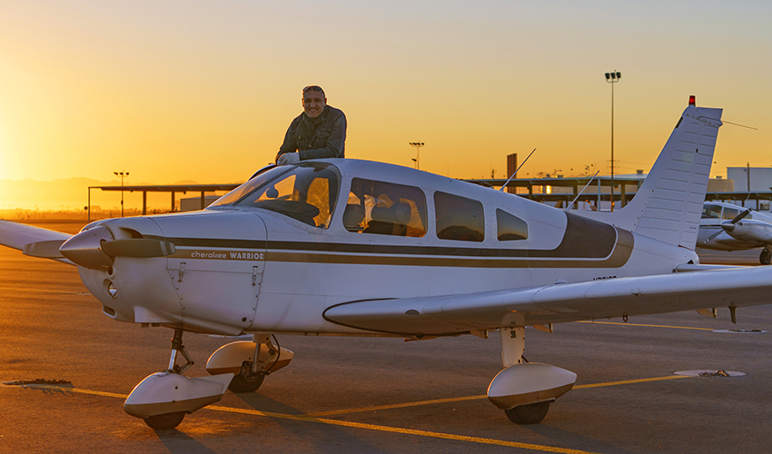 Jason poses by his airplane at an airport as the sun sets in the background.