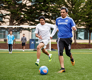 Two students chase a soccer ball on the field in front of student housing units, as two other players look on.