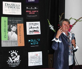 Photo of Catherine Kudlick with a microphone, standing next to a quilt made out of the late Paul Longmore's collection of disability related T-shirts.