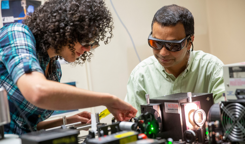 Two researchers wearing red goggles looking at a system of metal lab equipment