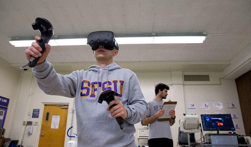 A student subject wearing a headset plays a virtual reality game in the Department of Kinesiology’s exercise lab while another student holds a clipboard and watches a computer monitor.