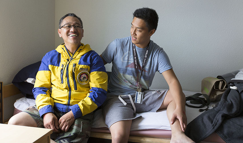 A student and family member sit on a bed inside a dorm room