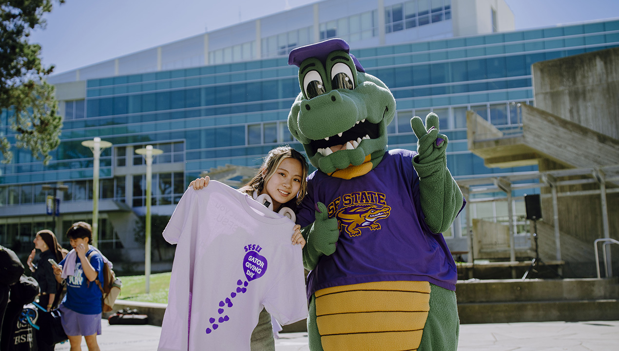 Student holding a lavendar Gator Giving Day shirt poses next to Alli Gator