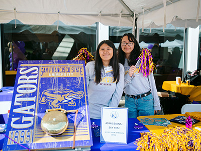 Two people pose for a picture while smiling and sanding at a table with a large bell on an SF State Gators sign and a sign that states Admissions Say Yes!