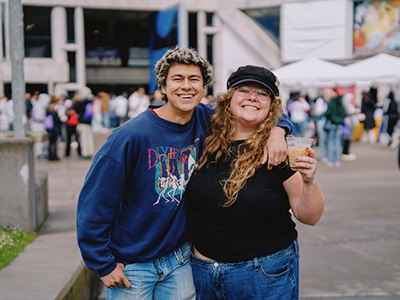Two people pose for a picture while smiling and standing on Malcolm X Plaza with one of them holding an iced coffee and wearing a hat