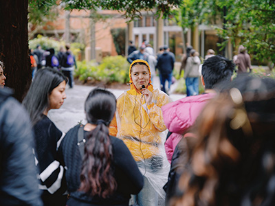 An SFSU student worker leads an outdoor campus tour surrounded by several people and while holding a microphone and wearing a yellow raincoat