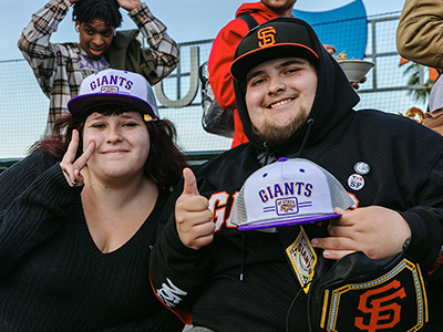 Two people pose for a photo while seated in the bleachers of Oracle Park with one of them flashing a peace sign and the other holding a co-branded SFSU/San Francisco Giants baseball cap